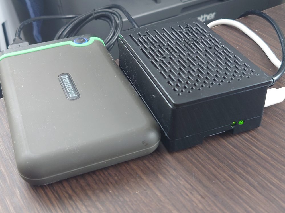 木製の机の上に置かれたRaspberry Pi 5。黒い放熱ケースに入り、緑のLEDが点灯している。左隣にはバックアップ用の外付けポータブルドライブが接続されている
